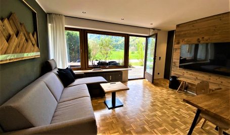 Heller Wohnraum mit Sofa, Holzelementen und Blick auf den grünen Außenbereich. Ferienwohnung Schleching