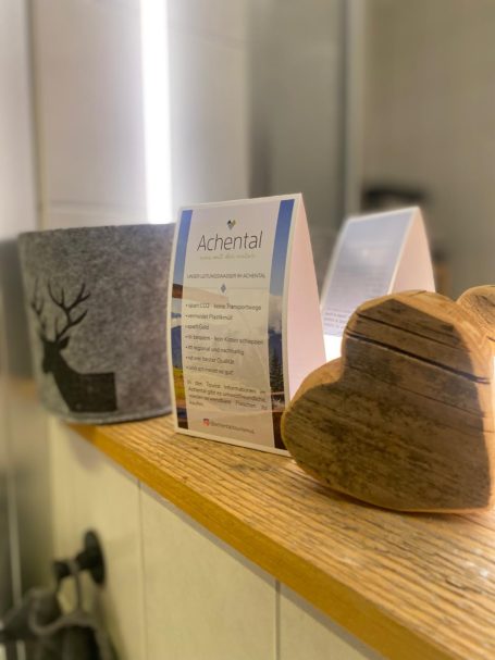 Eine graue Pflanzschale, eine herzförmige Holzdeko und ein Infozettel stehen auf einem Holzregal.