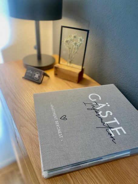 Graue Gästebuch mit dekorativer Tischlampe und Bildrahmen auf einem Holzregal.