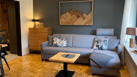 Wohnzimmer mit grauem Sofa, Holzmöbeln und einer Wanddekoration aus Holz. Ferienwohnung Schleching